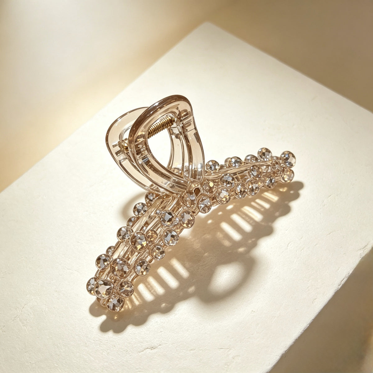 A clear plastic claw hair clip embellished with clear rhinestones, displayed on a textured cream-colored surface with warm, directional lighting.