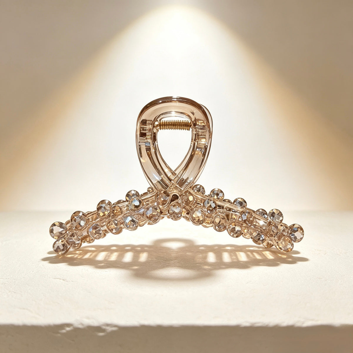 A clear acrylic claw hair clip embellished with sparkling clear rhinestones, displayed on a textured white surface under a warm spotlight.